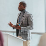 Speaker presenting at a professional seminar in a bright, modern conference room, engaging with a diverse audience. The concept of leadership, corporate training, and business communication is conveyed.